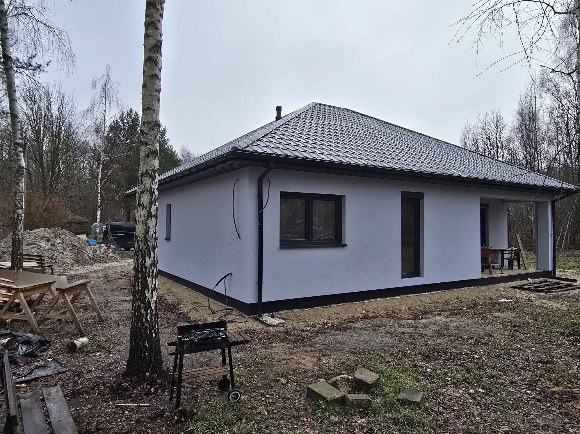 Gotowy, parterowy dom z szarą dachówką i białymi ścianami, widoczny z zewnątrz w jesiennej scenerii. Na pierwszym planie brzoza i grill.