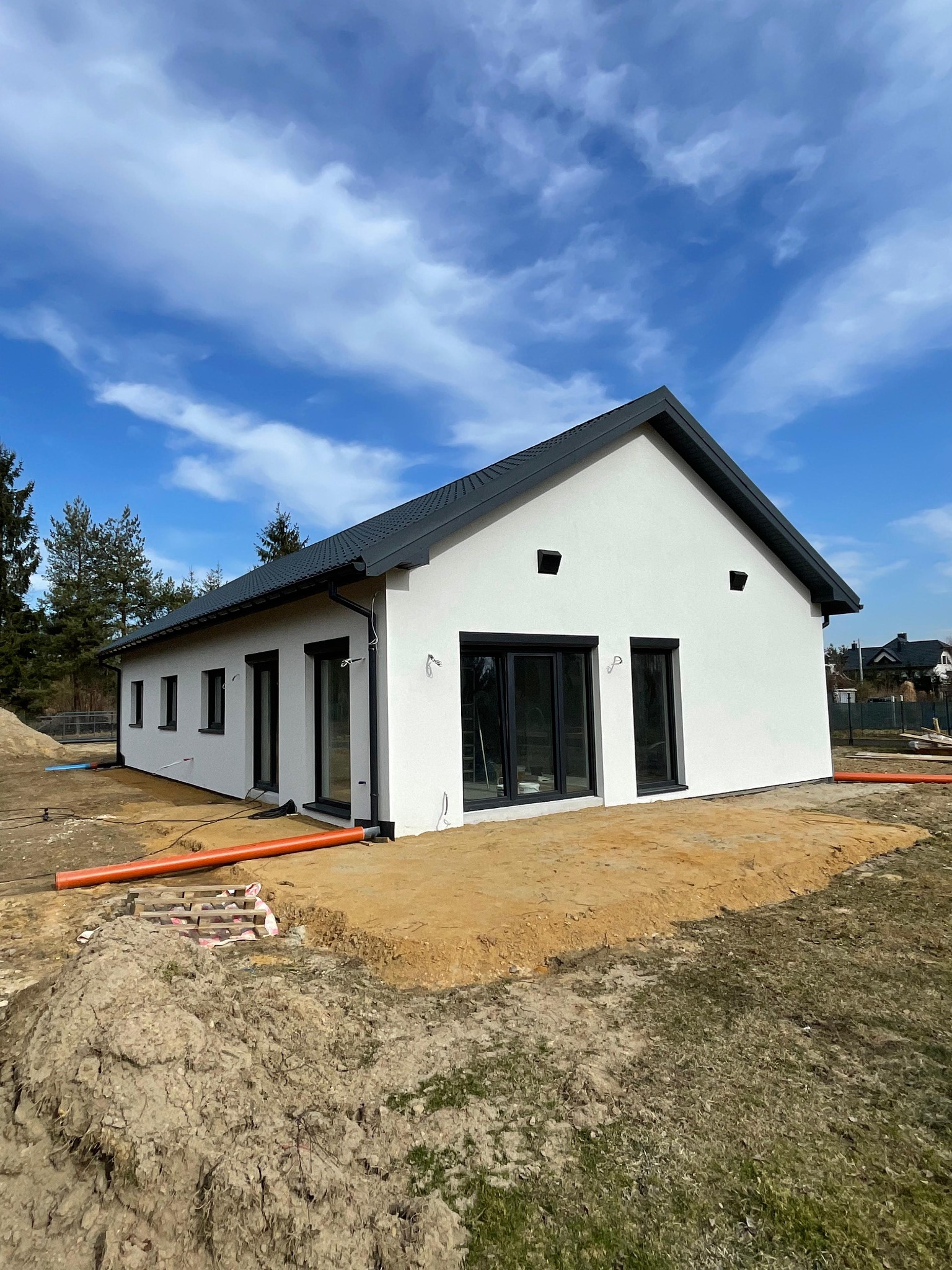 Biały parterowy dom z ciemnym dachem na działce w trakcie przygotowania, widoczne rury i ziemia. Minimalistyczna bryła, duże okna, błękitne niebo.