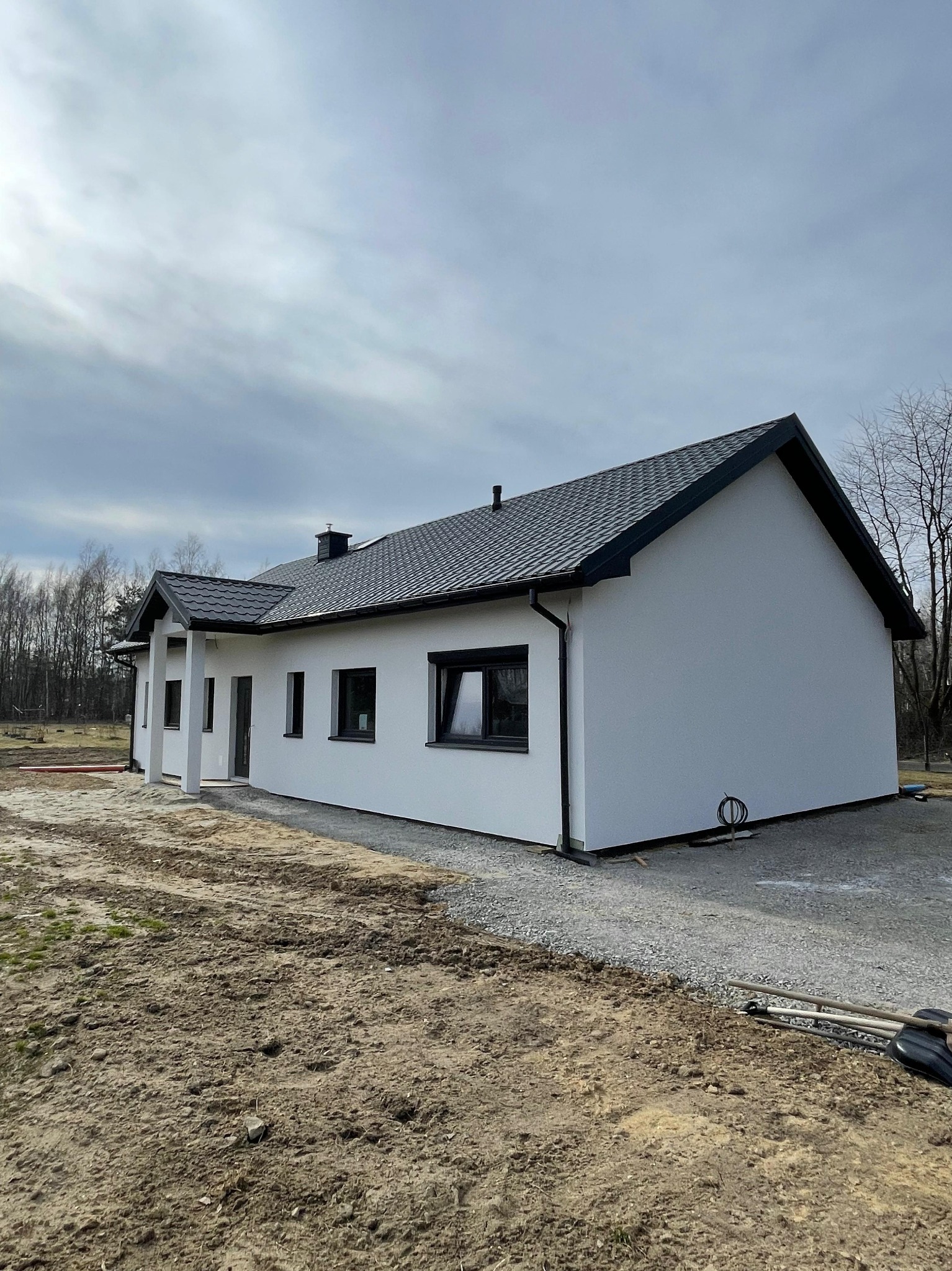 Gotowy, parterowy dom z białymi ścianami i ciemną dachówką, widoczny z boku. Na pierwszym planie ziemia i żwir, w tle drzewa bez liści. Pochmurne niebo.