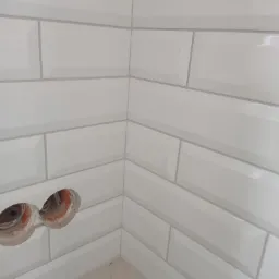 Świeżo ułożone, białe, błyszczące płytki typu metro w rogu łazienki, z widocznymi otworami na gniazdka elektryczne w trakcie montażu.