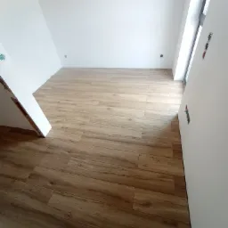 Ukończone układanie paneli podłogowych w nowym, jasnym pomieszczeniu z białymi ścianami i oknem, widoczne gniazdka elektryczne.