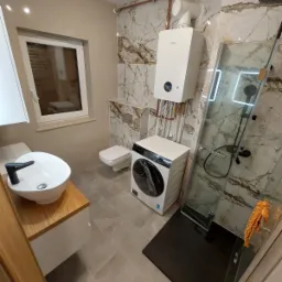 Nowoczesna łazienka z marmurowymi płytkami, umywalką nablatową na drewnianym blacie, pralką, kabiną prysznicową z czarnym brodzikiem i wiszącym piecem gazowym.