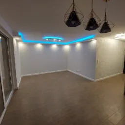 Puste, wykończone wnętrze pokoju z drewnianą podłogą, białymi ścianami z dekoracyjnym tynkiem, podwieszanym sufitem z niebieskim oświetleniem LED i trzema wiszącymi lampami w geometrycznych kloszach.