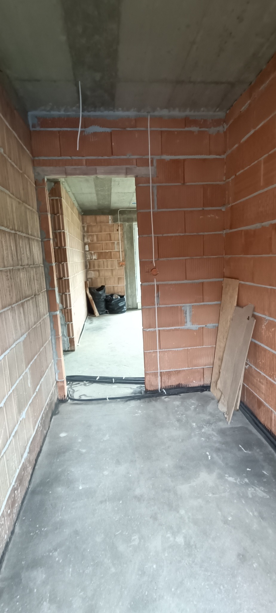 Wnętrze w stanie surowym z widocznymi przewodami elektrycznymi poprowadzonymi wzdłuż ścian i pod sufitem, kable ułożone przy podłodze, ściany z czerwonej cegły.