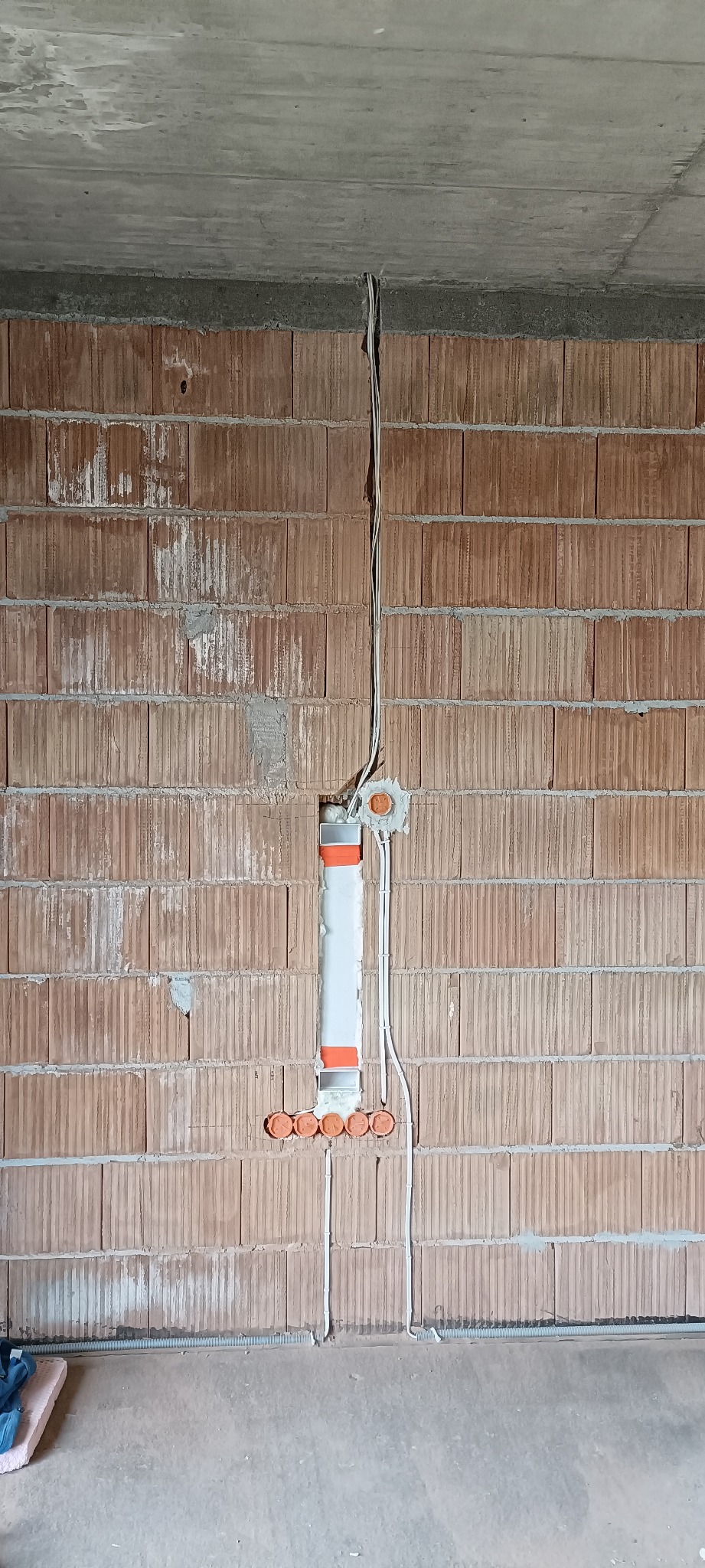 Surowa ściana z cegły ceramicznej z widoczną bruzdą pionową, w której umieszczono białą rurę instalacyjną z pomarańczowymi elementami mocującymi, oraz prowadzone przewody elektryczne w peszlu i bez...