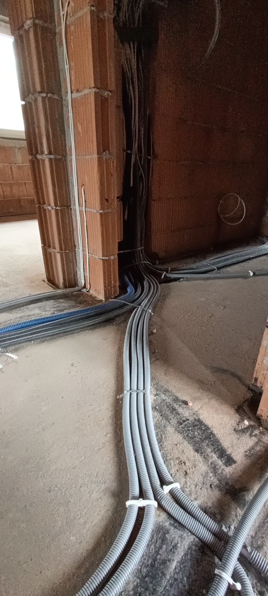 Wiązka szarych rur karbowanych prowadzona po podłodze w surowym stanie, przymocowana białymi opaskami zaciskowymi, w tle ściany z czerwonej cegły dziurawki i wystające kable.