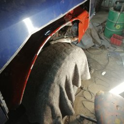 Sebastian Glinka - Fragment nadkola samochodu w trakcie naprawy blacharskiej, z widocznymi śladami szpachlowania i nałożoną warstwą podkładu w kolorze czerwonym. Przykryte kocem elementy.