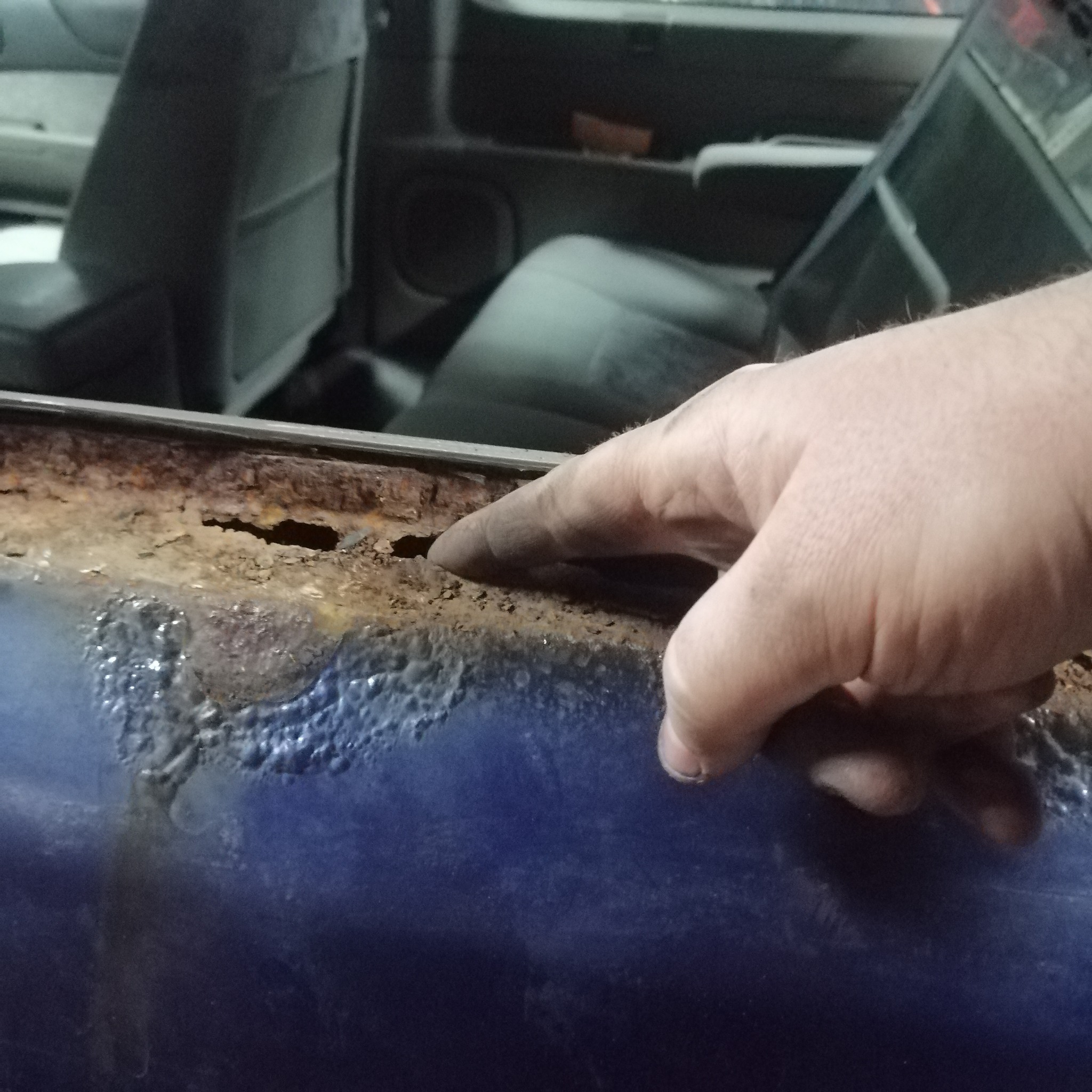 Korozja na karoserii auta, wskazana palcem. Widoczne ogniska rdzy i uszkodzenia lakieru. W tle wnętrze pojazdu. Detal z bliska, ukazujący stopień zniszczenia.