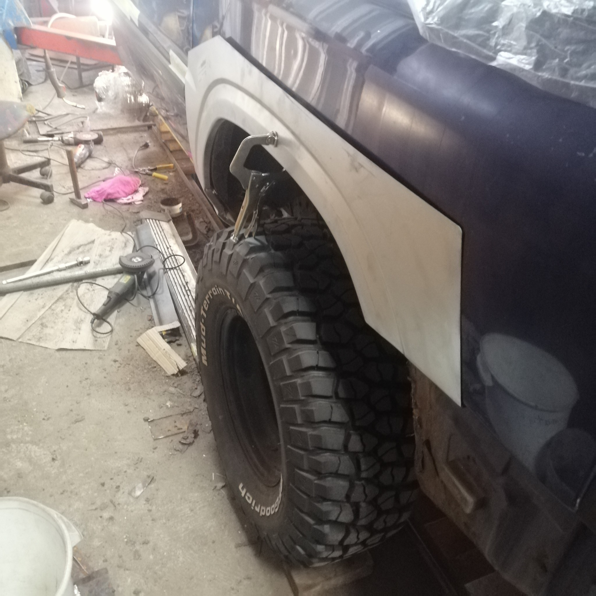 Błotnik w trakcie naprawy blacharskiej, z widocznym nowym elementem i narzędziami w warsztacie. Opona terenowa.