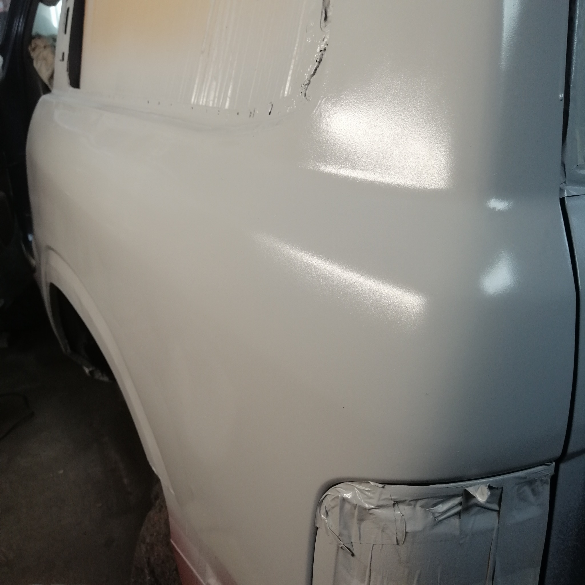 Świeżo polakierowany, jasnoszary bok samochodu dostawczego z widocznymi śladami taśmy maskującej. Błyszcząca powierzchnia odbija światło, ukazując gładką strukturę lakieru.