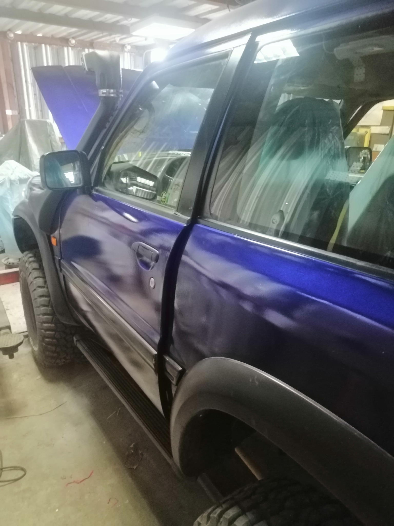 Niebieski samochód terenowy z otwartą maską w warsztacie. Widoczny snorkel, opony terenowe i folia ochronna na siedzeniach. Detal po lakierowaniu.