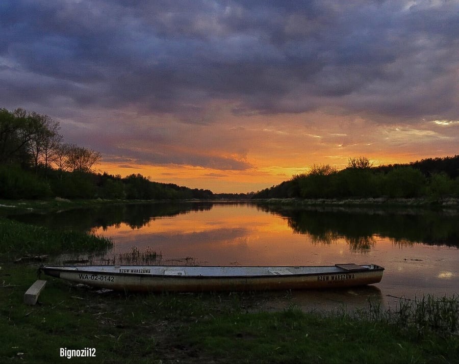 Spokojny krajobraz rzeki o zmierzchu z łodzią przy brzegu, odbicie zachodzącego słońca w wodzie, ciemne chmury nad horyzontem.