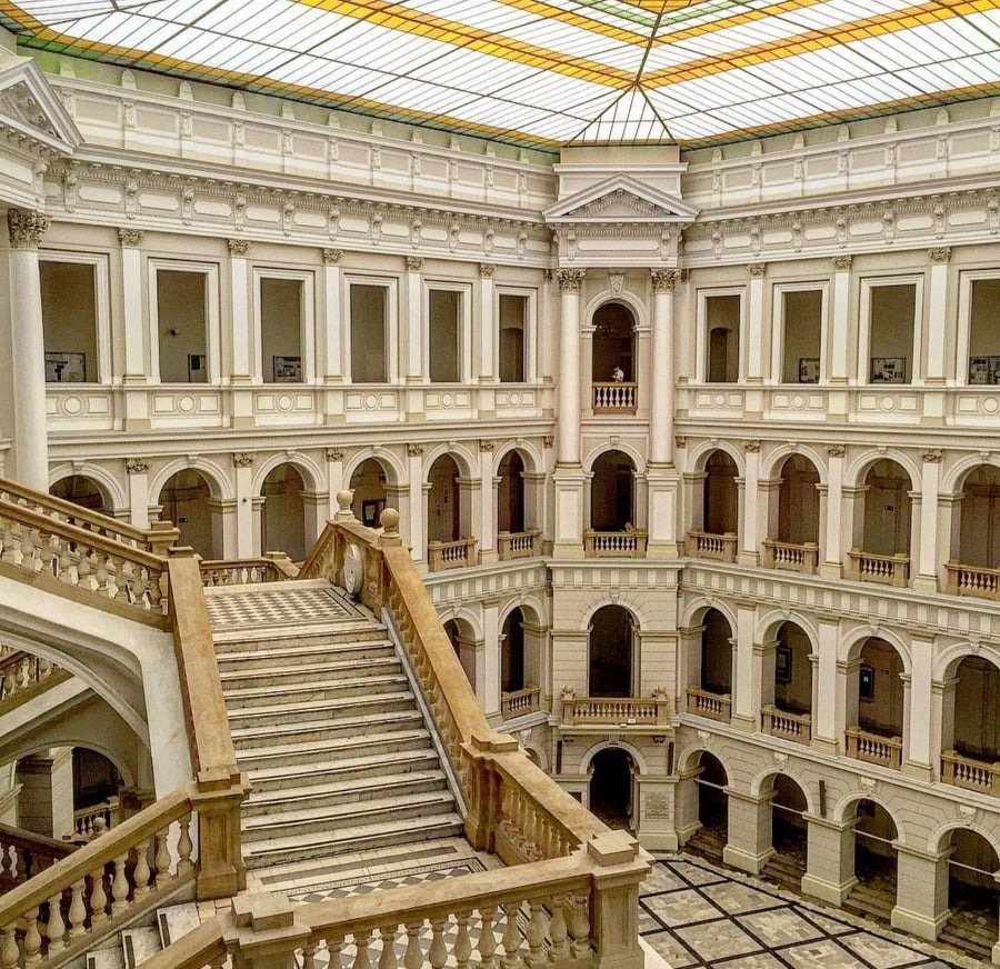 Wnętrze monumentalnego budynku z jasnego kamienia z wielopoziomowymi galeriami, łukami i schodami z marmurową szachownicą. Przeszklony sufit w geometryczne wzory w odcieniach żółci i bieli.