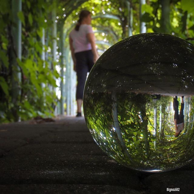 Szklana kula na brukowanej ścieżce odbija zielony tunel roślinny z idącą w oddali kobietą.