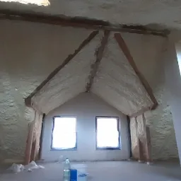 Poddasze po natrysku pianki izolacyjnej na skosach dachu i ścianach, widoczne drewniane elementy konstrukcyjne, dwa okna dachowe i butelki z wodą na podłodze.