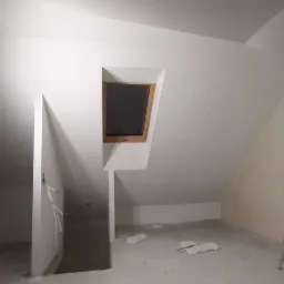 Poddasze po odświeżeniu, z oknami dachowymi, ścianami w kolorze białym, z widoczną drabiną i lampą roboczą na statywie.