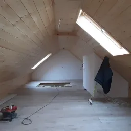 Poddasze z drewnianą boazerią na ścianach i suficie, widoczne okna dachowe, fragment podłogi z panelami i narzędzie pracy w rogu, odzież robocza zawieszona na ścianie.