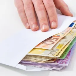 Dłoń wsuwa plik banknotów euro o różnych nominałach (50, 100, 200, 500) do białej koperty na jasnym tle.