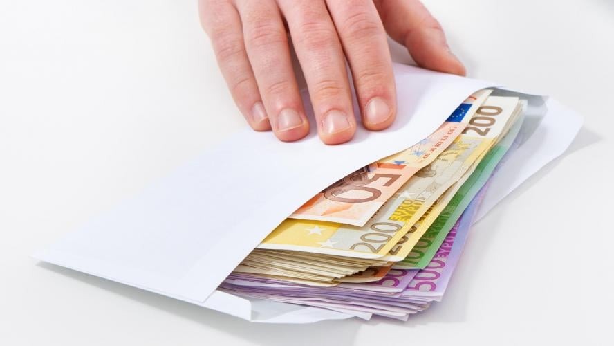 Dłoń wsuwa plik banknotów euro o różnych nominałach (50, 100, 200, 500) do białej koperty na jasnym tle.