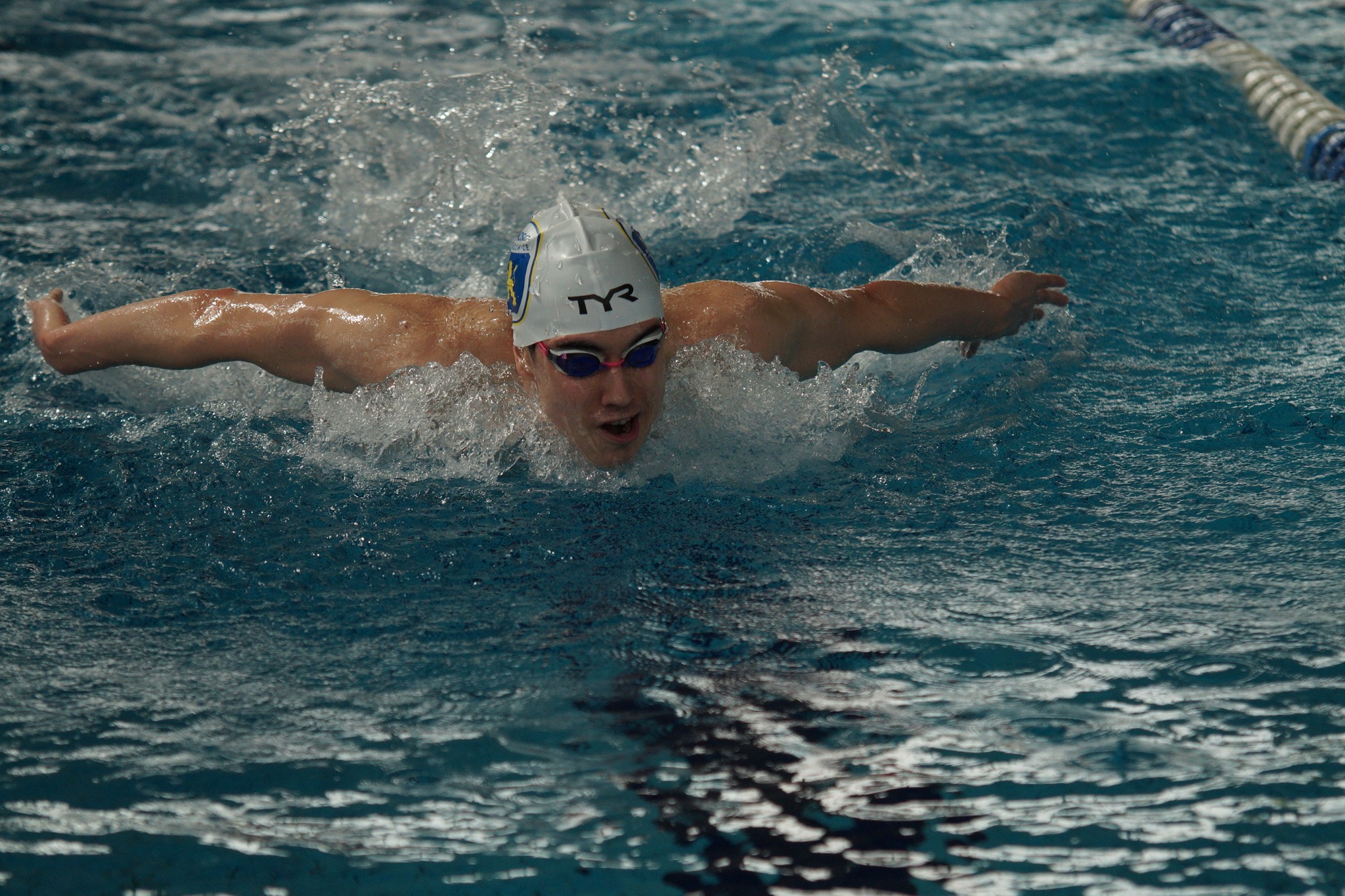 Pływak w czepku i okularach, z otwartymi ustami, płynie stylem motylkowym w basenie, z widocznymi falami i pluskiem wody.