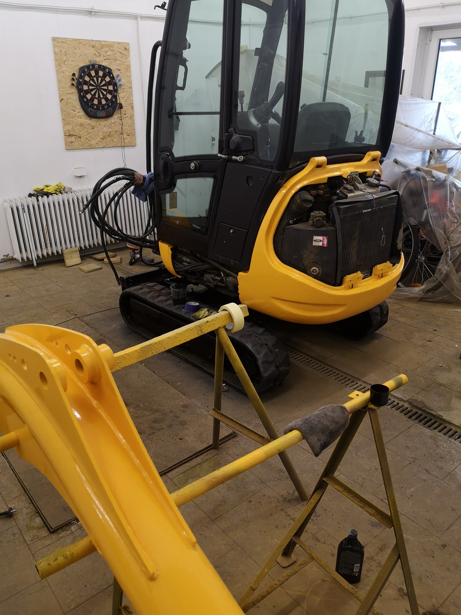 Żółty mini-koparka w trakcie renowacji w warsztacie, z odłączonym ramieniem na kozłach malarskich, widoczna tarcza do darta w tle.