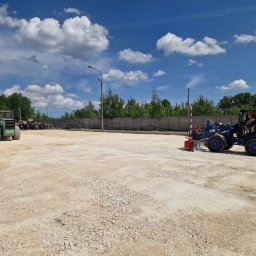 EKOPRINŻ SP. Z O.O. - Niebieska mini ładowarka CAT 903D z operatorem wyrównuje teren pokryty drobnym kruszywem, w tle zielony walec i betonowy płot w słoneczny dzień.