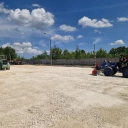 Niebieska mini ładowarka CAT 903D z operatorem wyrównuje teren pokryty drobnym kruszywem, w tle zielony walec i betonowy płot w słoneczny dzień.