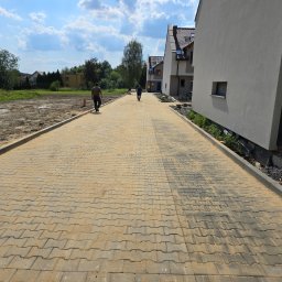 EKOPRINŻ SP. Z O.O. - Żółta kostka brukowa ułożona na długiej ścieżce między nowymi domami, dwóch mężczyzn idzie w oddali, z lewej strony widać przygotowany teren pod budowę.