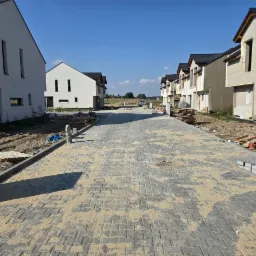Ulica z nową kostką brukową w Dąbrowie Górniczej, pomiędzy nowo budowanymi domami jednorodzinnymi, piasek na kostce, w tle widoczny sprzęt budowlany.