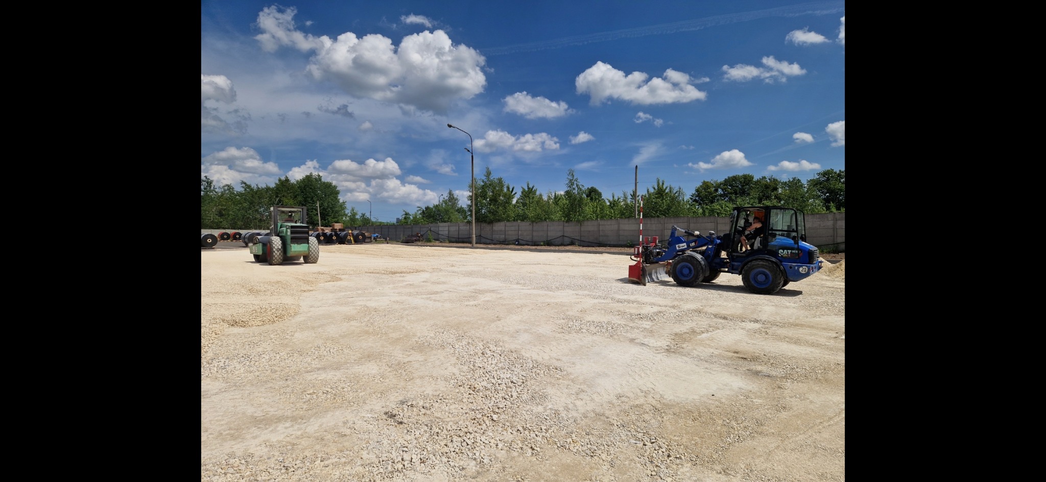 Niebieska mini ładowarka CAT 903D z operatorem wyrównuje teren pokryty drobnym kruszywem, w tle zielony walec i betonowy płot w słoneczny dzień.