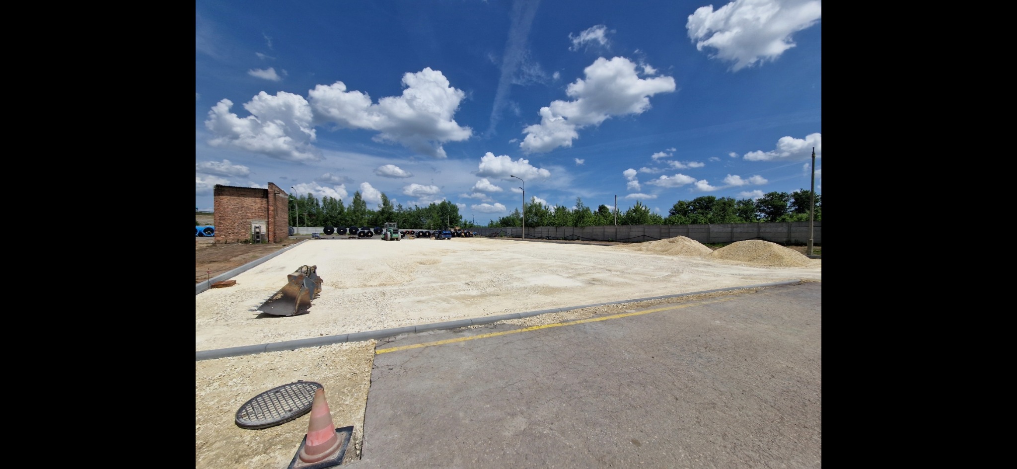 Przygotowany teren pod budowę z widocznymi maszynami budowlanymi w tle, hałdami piasku i fragmentem starego budynku. Ujęcie z poziomu gruntu.