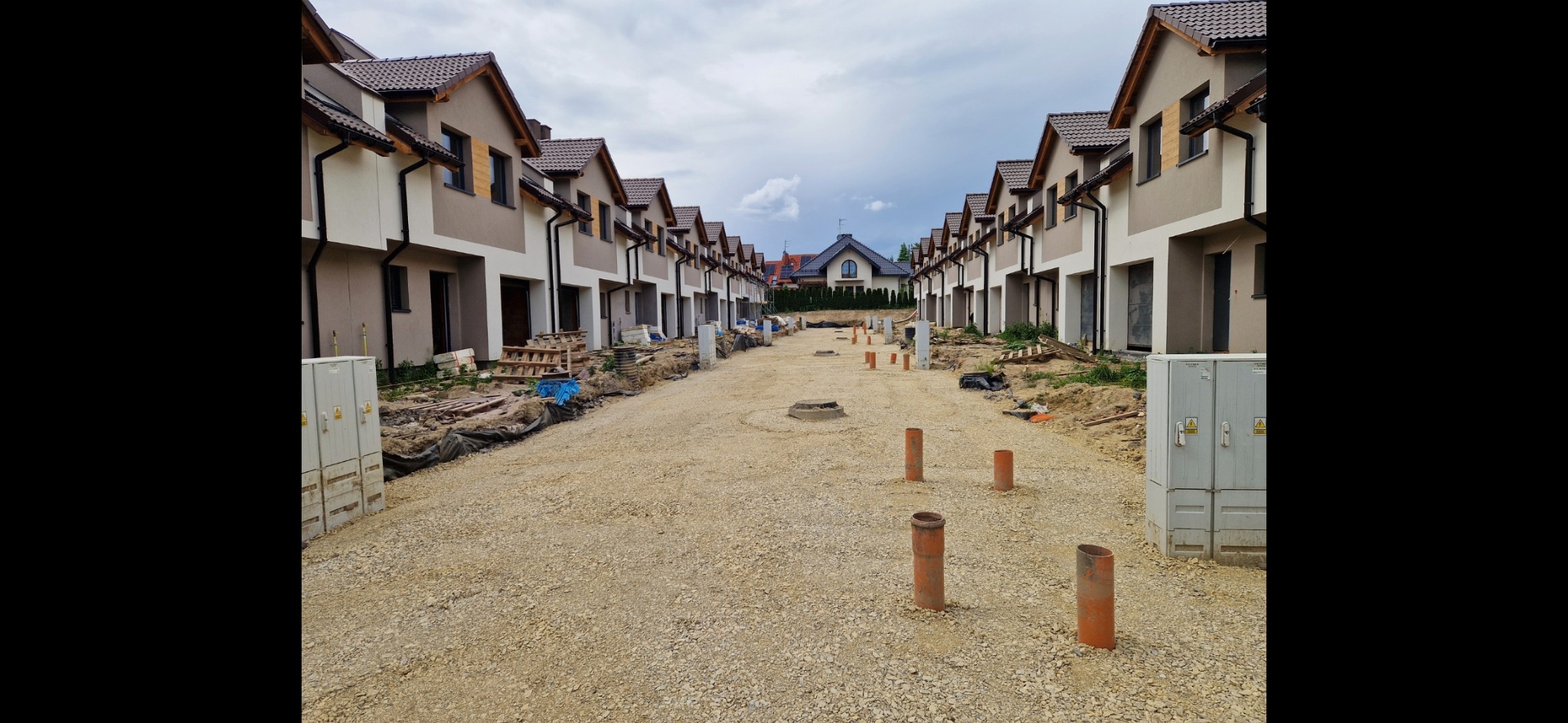 Ulica w trakcie budowy osiedla domów jednorodzinnych, widoczne rury kanalizacyjne wystające z gruntu, przygotowanie pod nawierzchnię drogi.