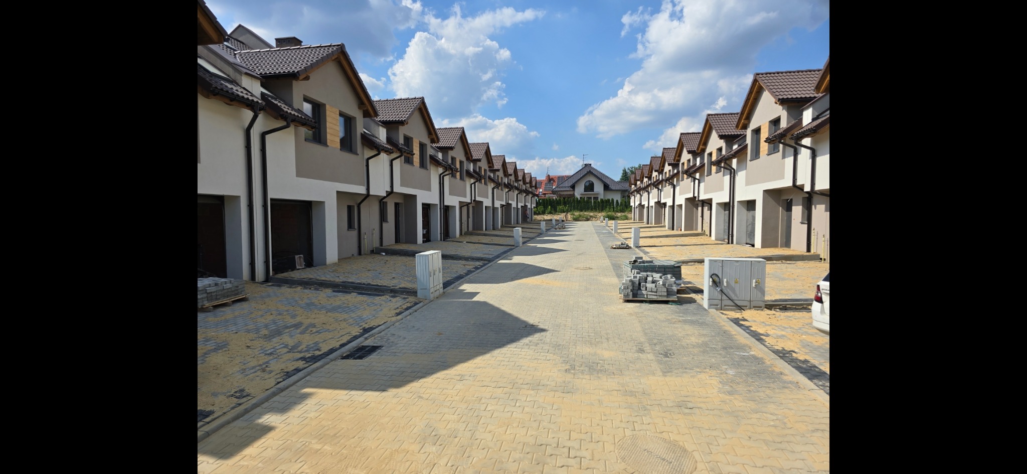 Nowo wybudowane osiedle domów szeregowych z częściowo ułożoną kostką brukową na podjeździe, widoczne palety z kostką i skrzynka elektryczna, błękitne niebo w tle.
