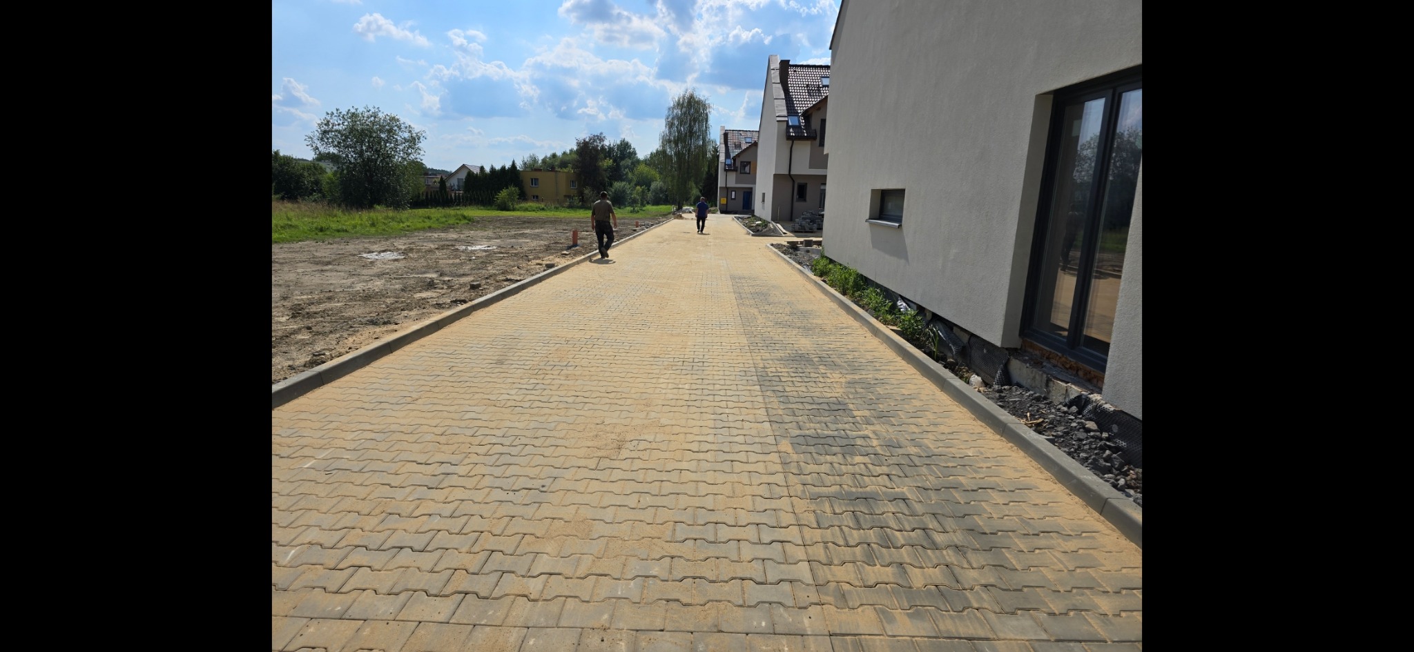 Żółta kostka brukowa ułożona na długiej ścieżce między nowymi domami, dwóch mężczyzn idzie w oddali, z lewej strony widać przygotowany teren pod budowę.