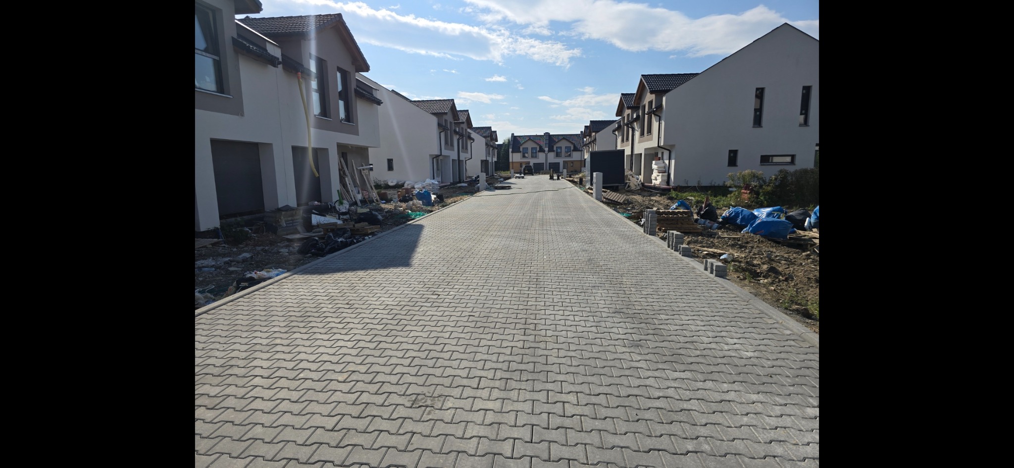 Ulica osiedlowa wyłożona szarą kostką brukową, nowe domy w tle, widoczne materiały budowlane i pozostałości po budowie.