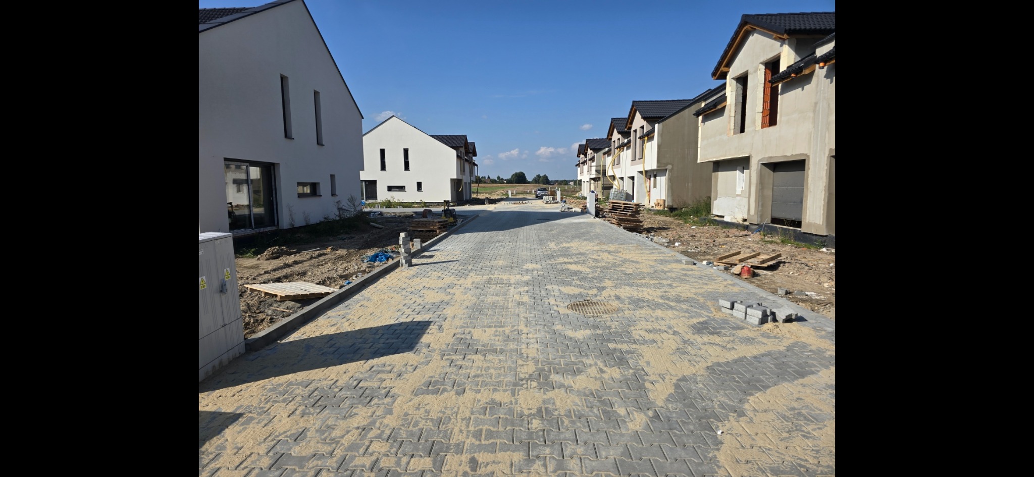Ulica z nową kostką brukową w Dąbrowie Górniczej, pomiędzy nowo budowanymi domami jednorodzinnymi, piasek na kostce, w tle widoczny sprzęt budowlany.