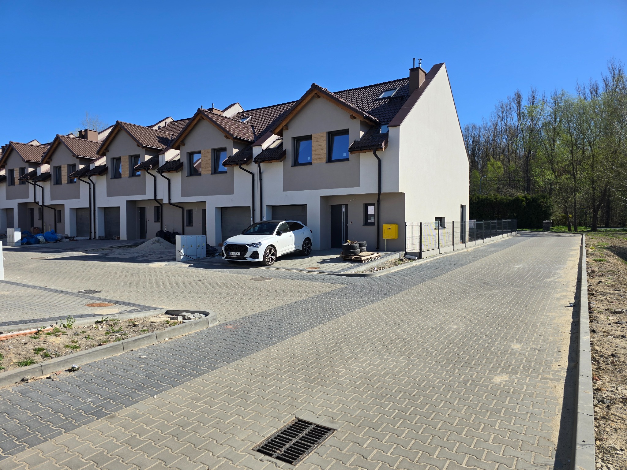 Nowe osiedle domów szeregowych z kostki brukowej, biały samochód zaparkowany przed jednym z budynków, widoczny fragment drogi w budowie.