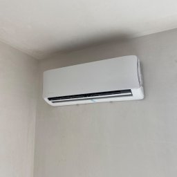 Andrzej Stoga Asklima - Nowoczesny, biały klimatyzator marki Rotenso zamontowany na szarej, surowej ścianie w rogu pomieszczenia. Widoczna niebieska taśma zabezpieczająca na urządzeniu.