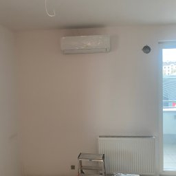 Andrzej Stoga Asklima - Nowo zamontowana jednostka klimatyzacji, jeszcze w folii ochronnej, na białej ścianie w pokoju z grzejnikiem i drabinką, widoczne ślady prac wykończeniowych.