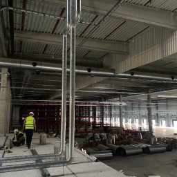 Andrzej Stoga Asklima - Montaż pionowych rur wentylacyjnych w hali magazynowej, widoczni pracownicy w kamizelkach odblaskowych i kaskach, surowe wykończenie wnętrza, widoczne elementy konstrukcyjne budynku i materiały...