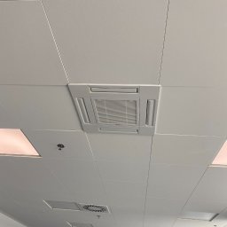 Andrzej Stoga Asklima - Kasetonowa klimatyzacja sufitowa w biurze, widok z dołu, kratka wentylacyjna, czujnik dymu i oświetlenie w tle.