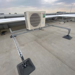 Andrzej Stoga Asklima - Klimatyzator Mitsubishi Electric zamontowany na metalowej konstrukcji wsporczej na płaskim dachu budynku przemysłowego, widoczne rury wentylacyjne w tle i pochmurne niebo.