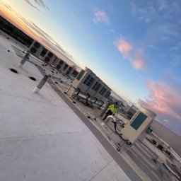 Andrzej Stoga Asklima - Praca na dachu płaskim przy instalacji klimatyzatorów i wentylacji, widoczny pracownik w kasku i kamizelce odblaskowej, rury wentylacyjne, tło z zachodem słońca.