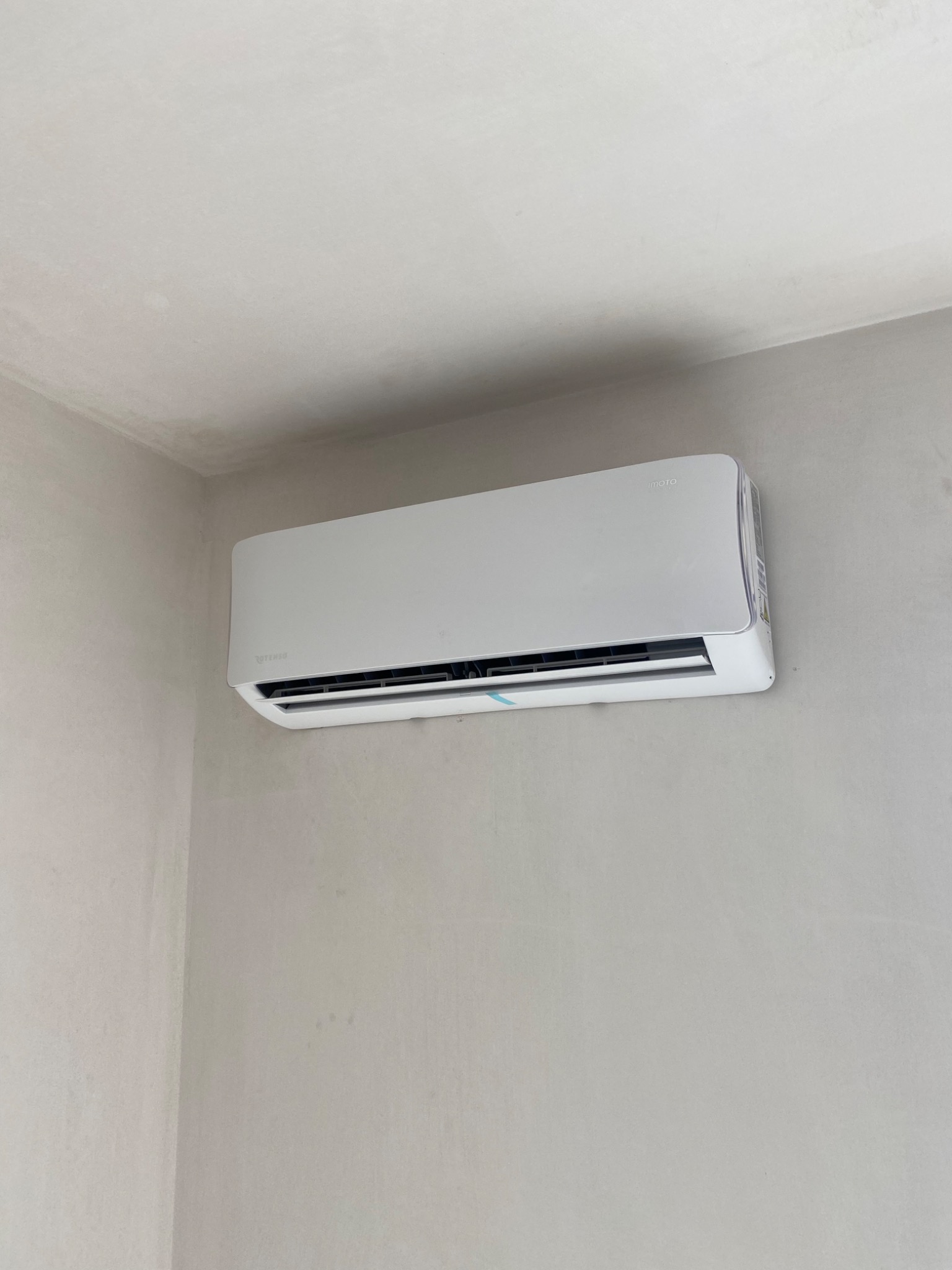 Nowoczesny, biały klimatyzator marki Rotenso zamontowany na szarej, surowej ścianie w rogu pomieszczenia. Widoczna niebieska taśma zabezpieczająca na urządzeniu.