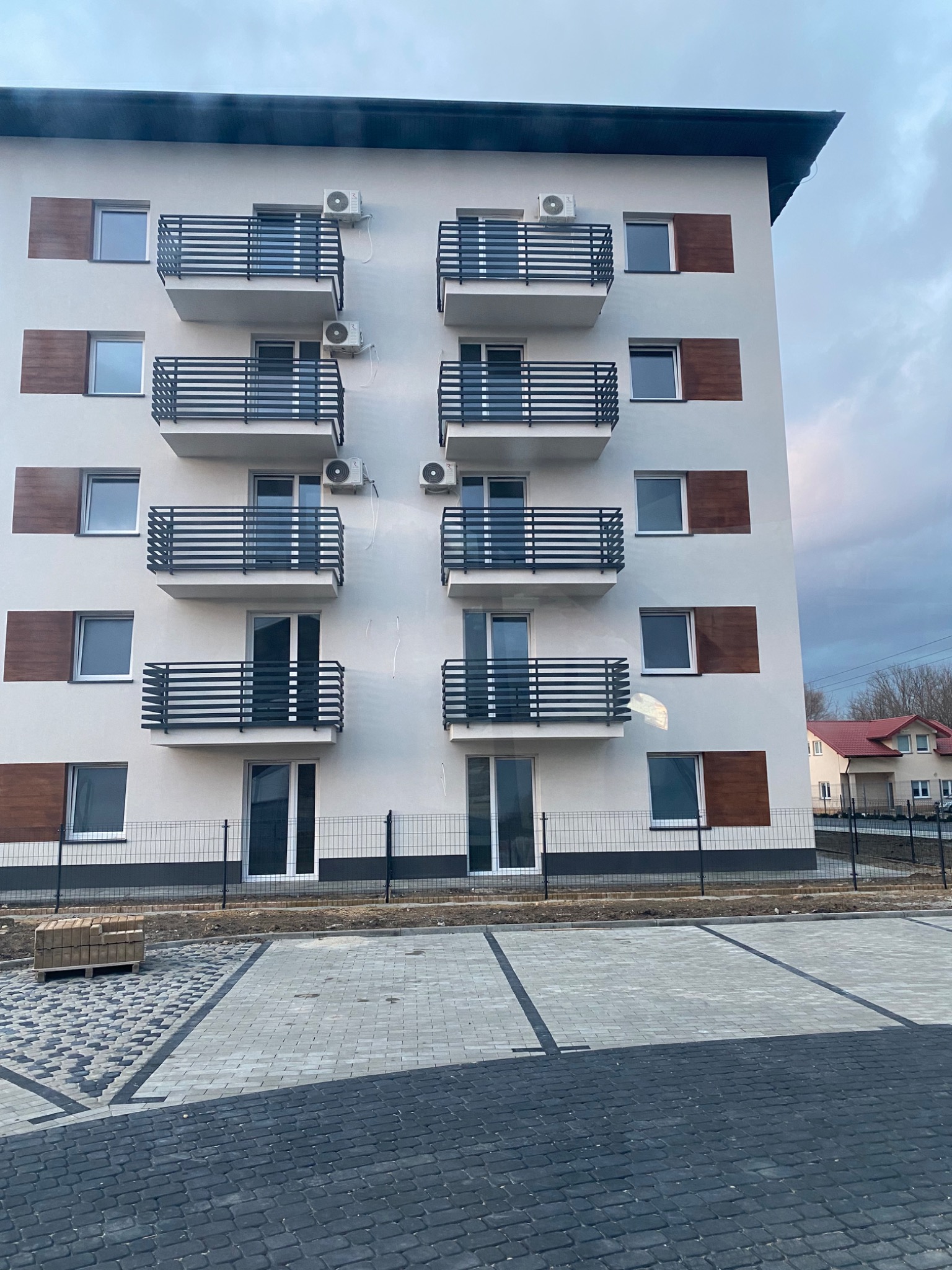 Biały budynek wielorodzinny z balkonami i zamontowanymi klimatyzatorami na elewacji, widok z poziomu parkingu wyłożonego kostką brukową.