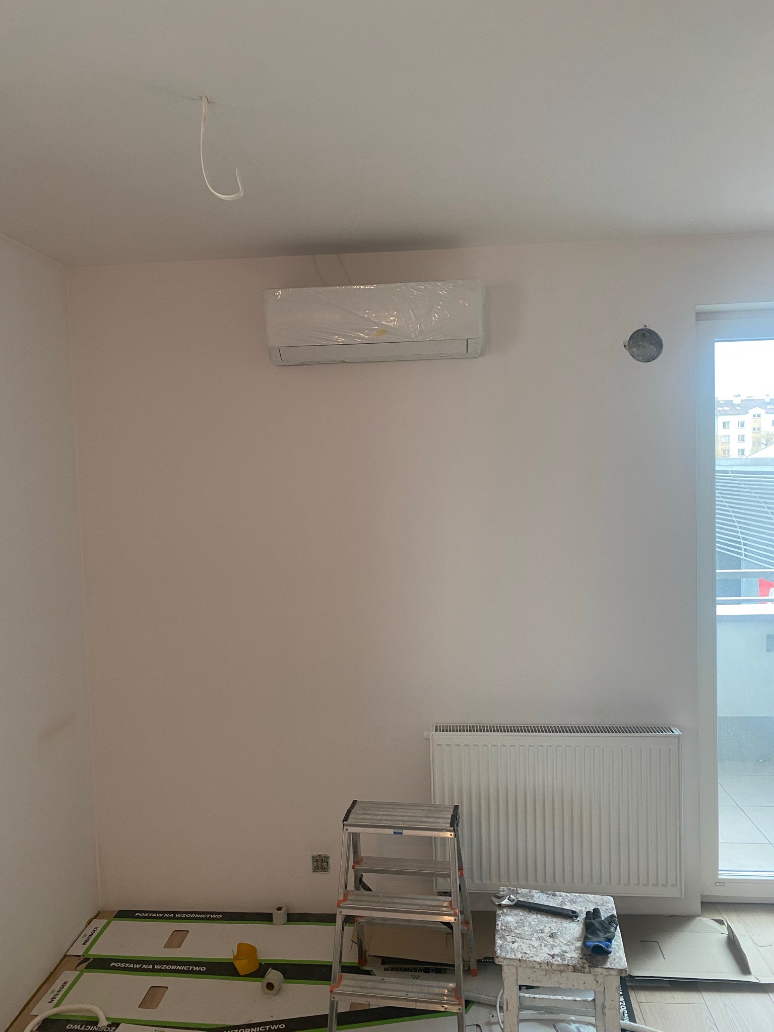 Nowo zamontowana jednostka klimatyzacji, jeszcze w folii ochronnej, na białej ścianie w pokoju z grzejnikiem i drabinką, widoczne ślady prac wykończeniowych.