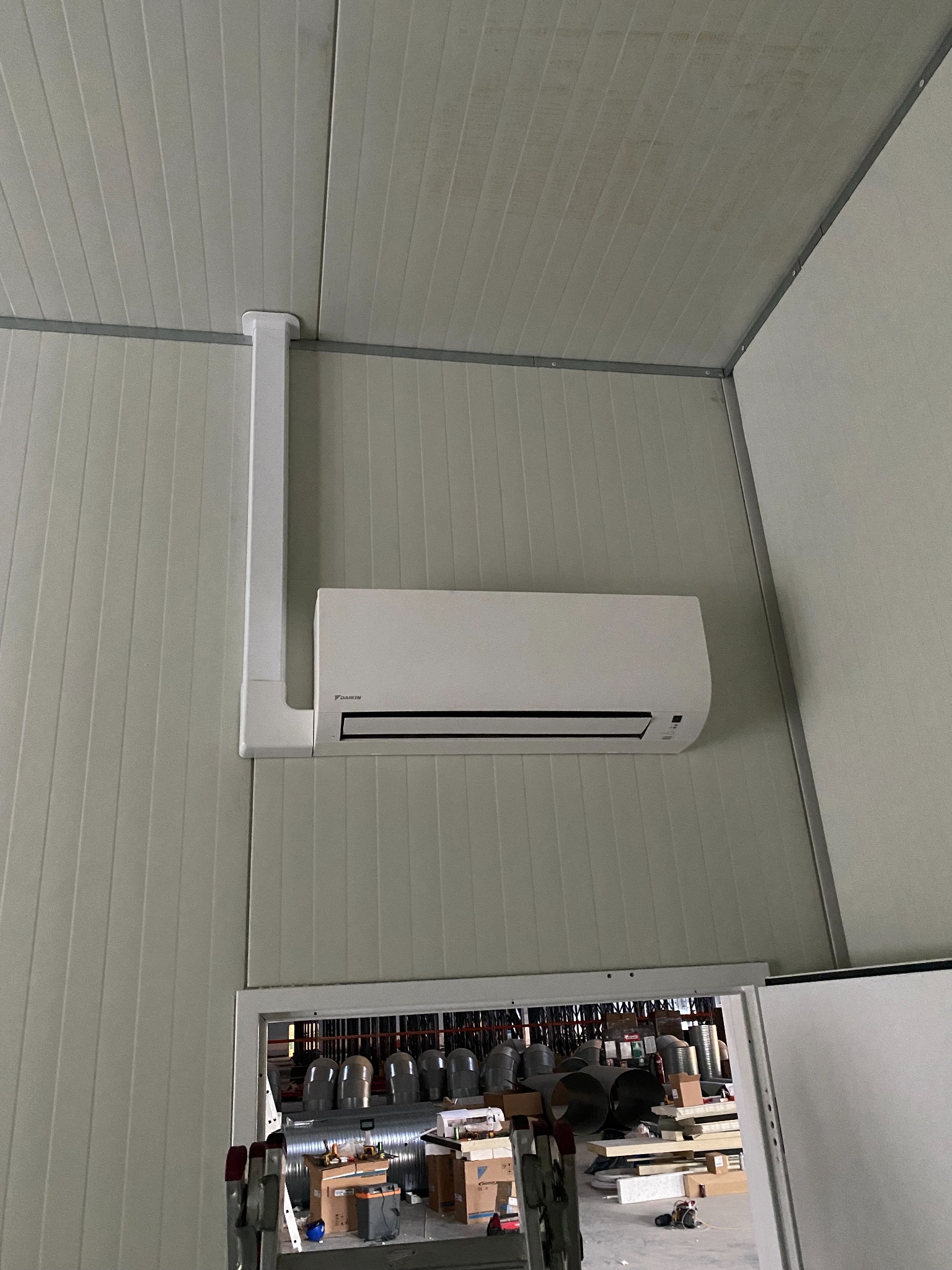 Zamontowany klimatyzator Daikin na ścianie z paneli, widoczne rury instalacyjne i fragment pomieszczenia z elementami wentylacji przez otwarte drzwi.