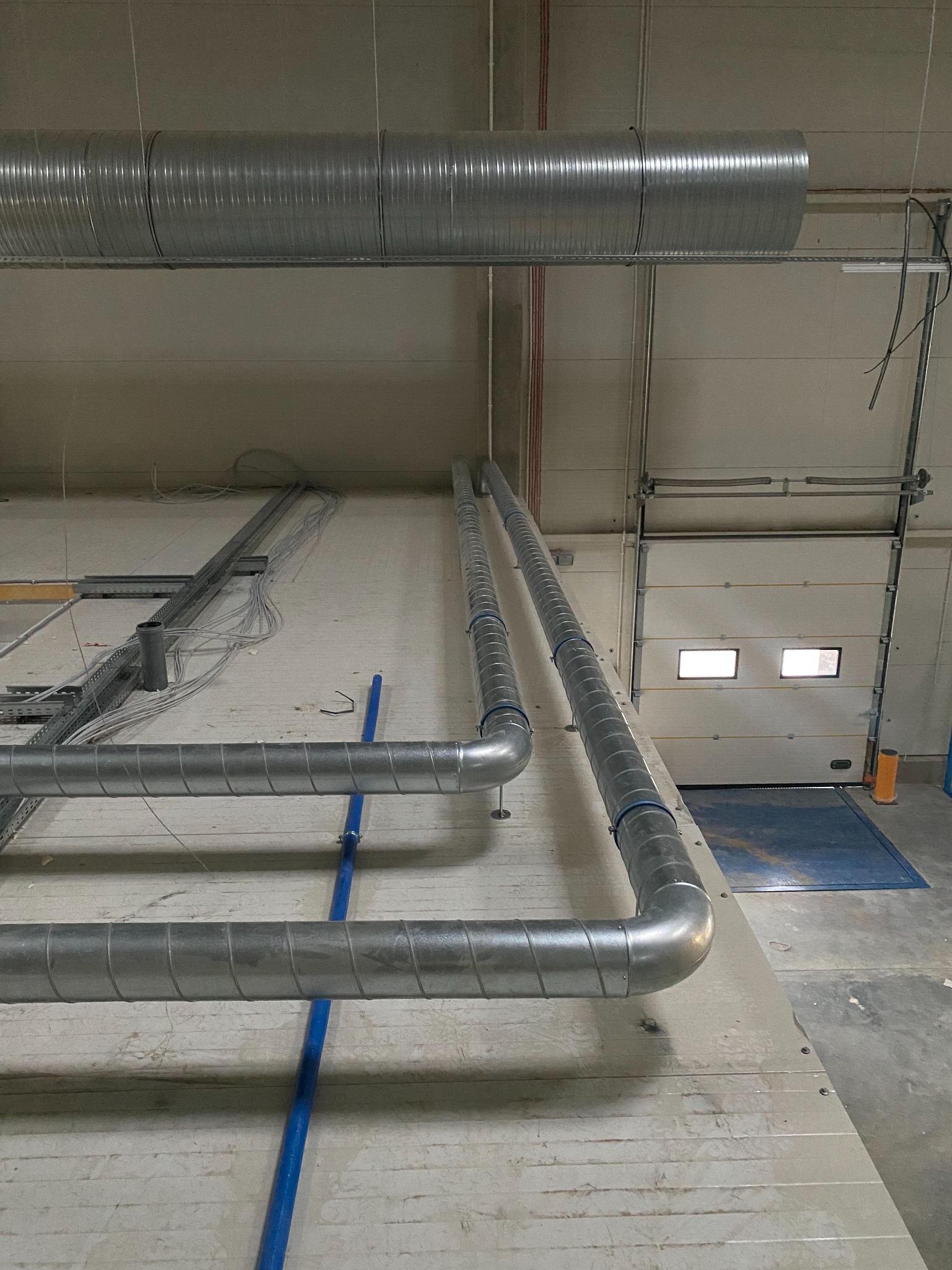 Metalowe rury wentylacyjne, spiralne, biegnące pod sufitem w hali przemysłowej, z widocznymi elementami mocującymi i instalacją elektryczną. W tle brama garażowa z oknami.