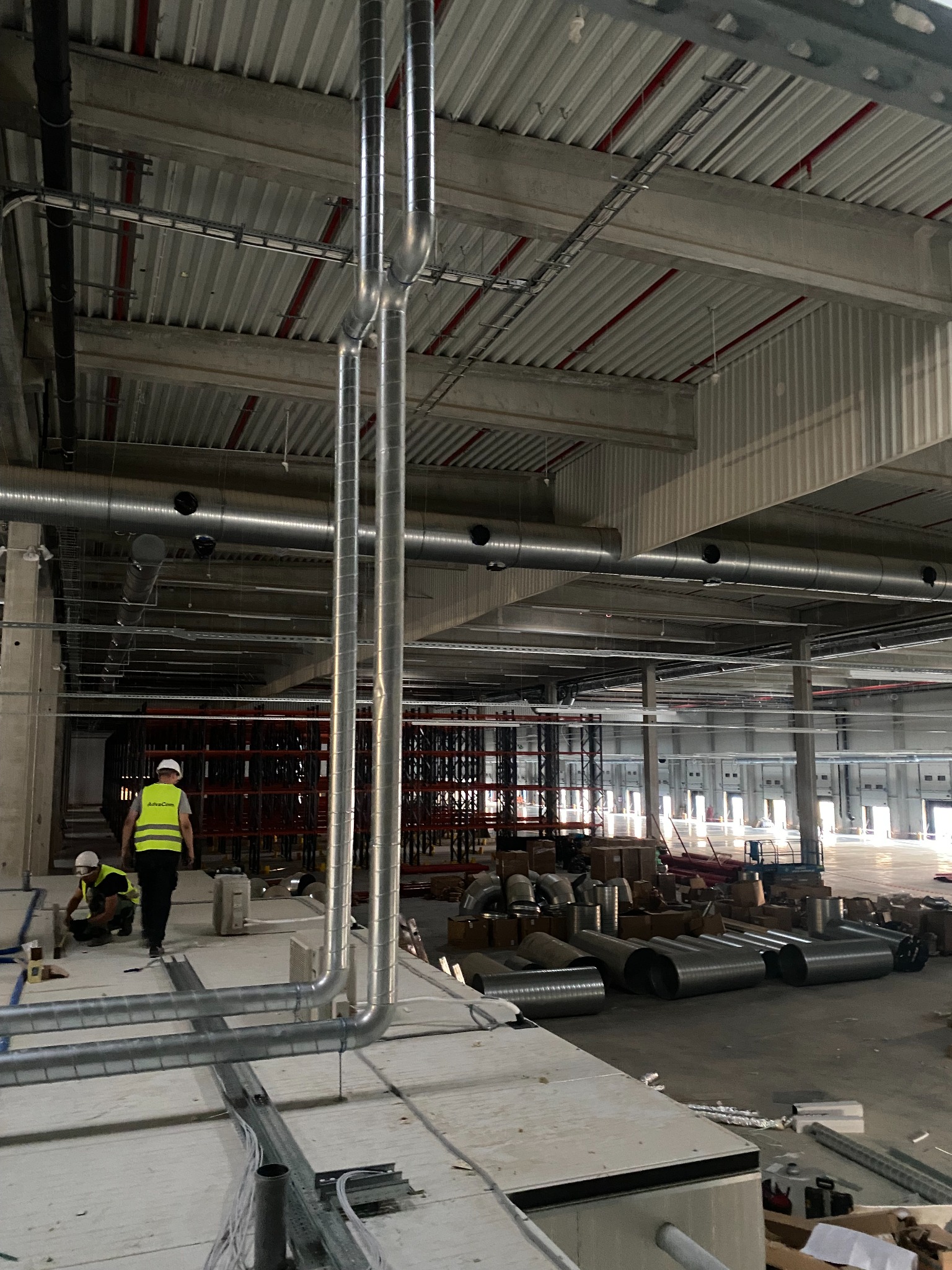 Montaż pionowych rur wentylacyjnych w hali magazynowej, widoczni pracownicy w kamizelkach odblaskowych i kaskach, surowe wykończenie wnętrza, widoczne elementy konstrukcyjne budynku i materiały...
