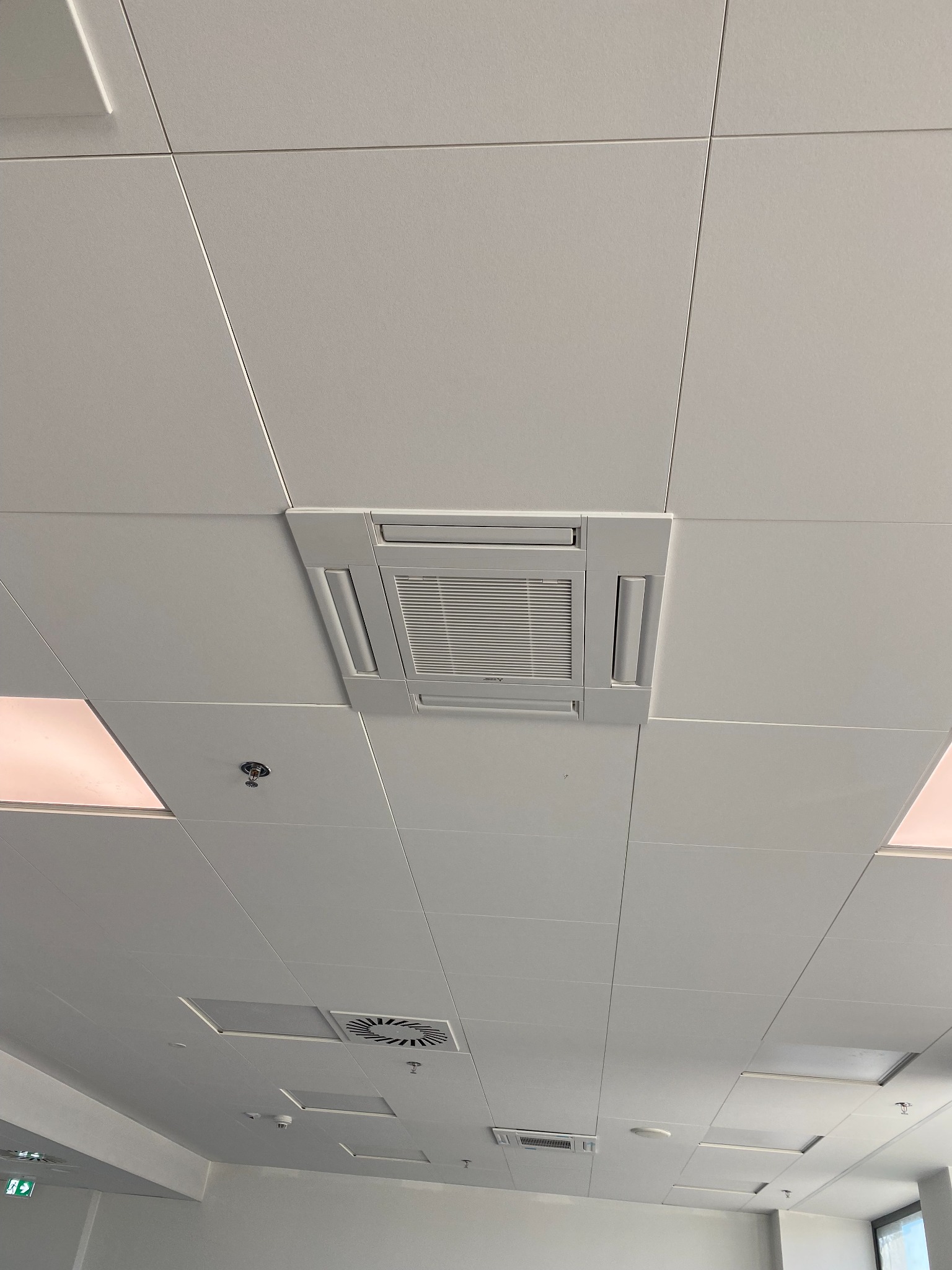 Kasetonowa klimatyzacja sufitowa w biurze, widok z dołu, kratka wentylacyjna, czujnik dymu i oświetlenie w tle.